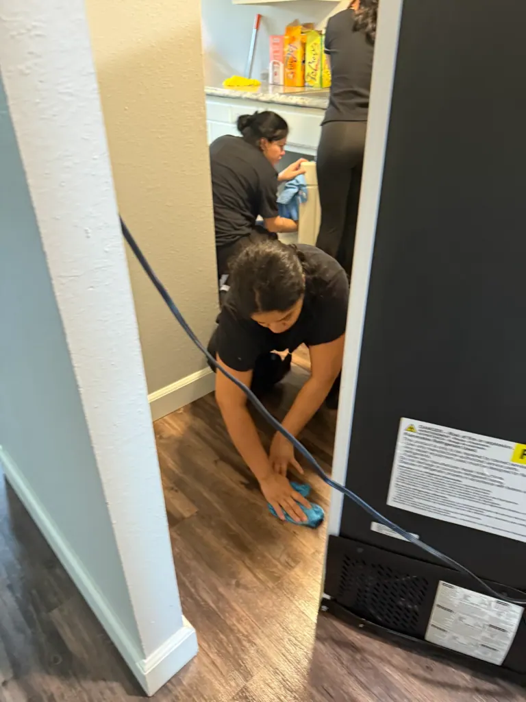 A Reina's Cleaning cleaner wiping the floor behind a pulled-out refrigerator